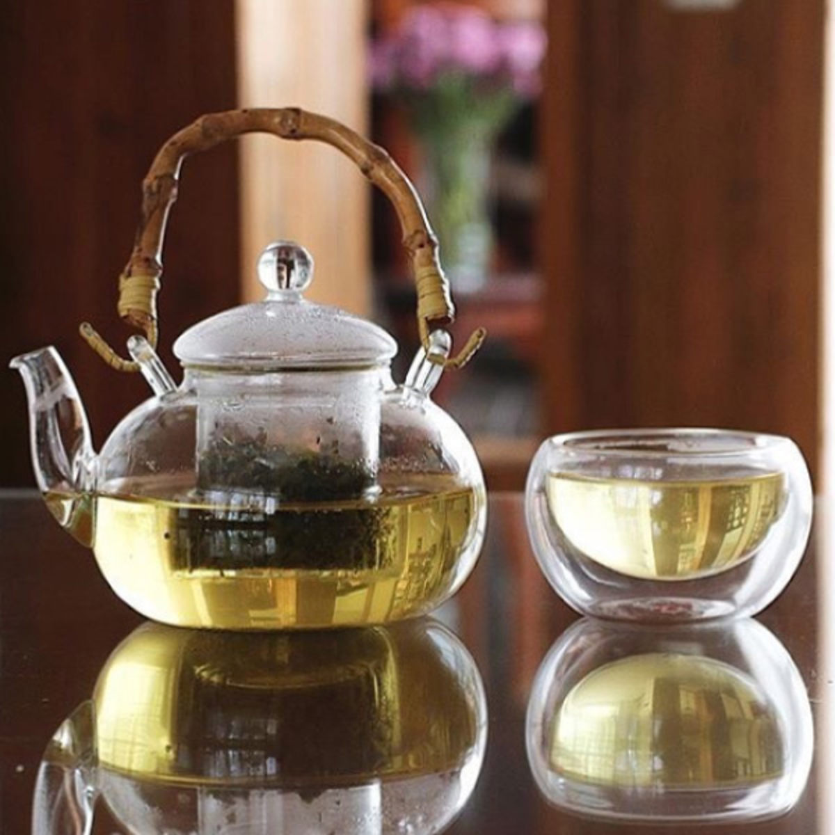 Glass teapot with bamboo handle filled with green tea on a table next to a matching glass teacup.