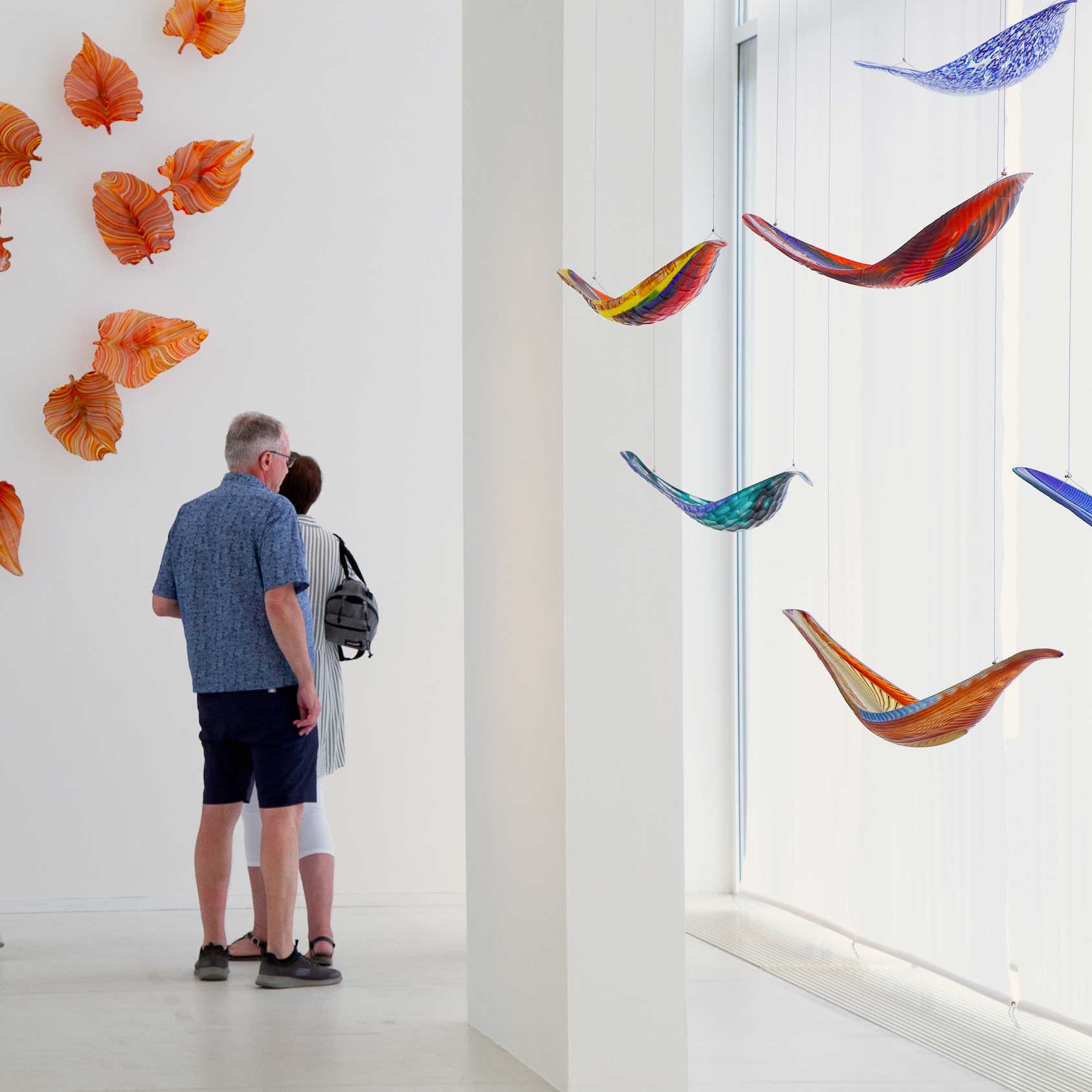 Two people admire colorful glass sculpture installation in a modern gallery exhibition.