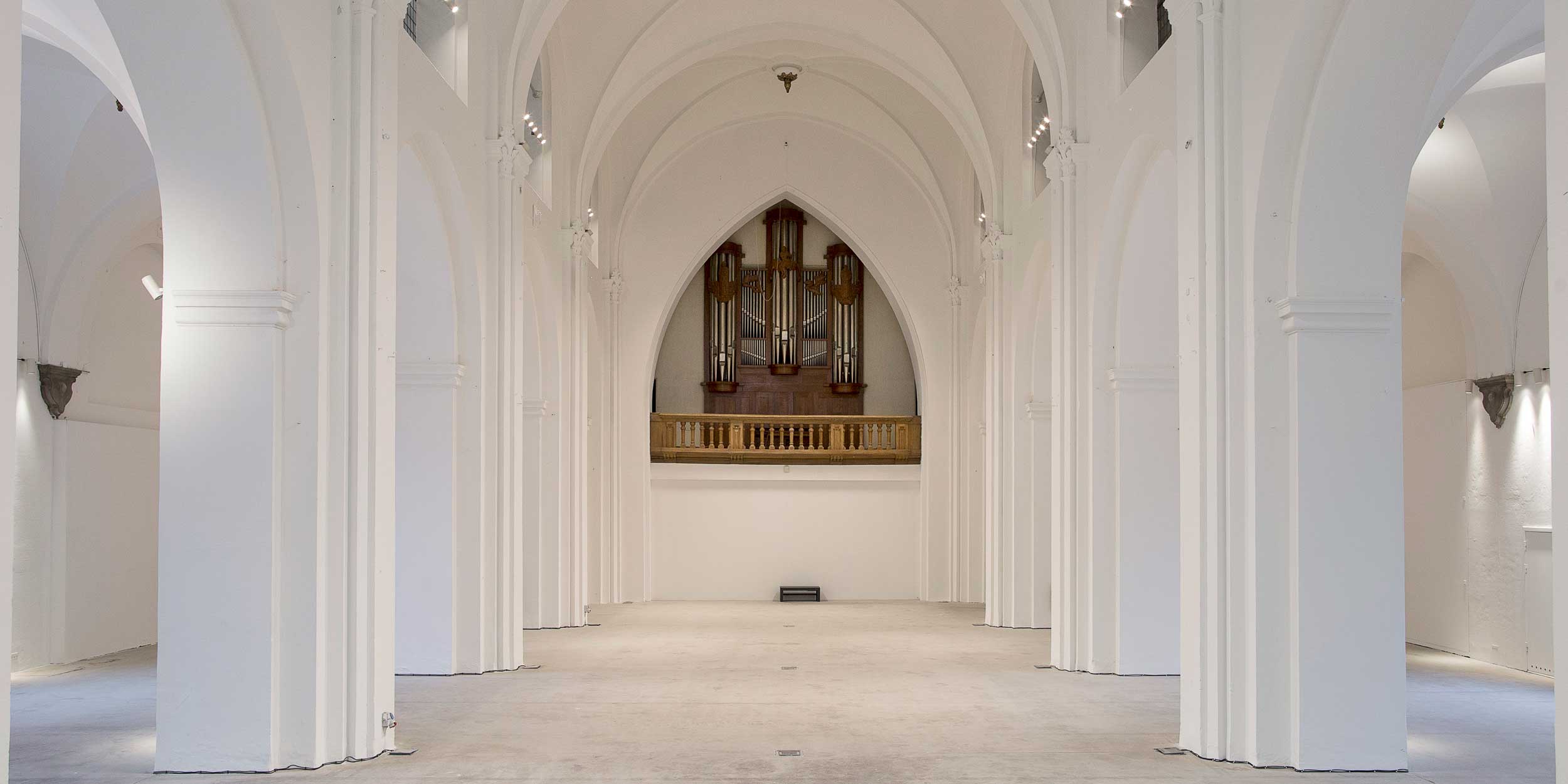 Spacious white church interior with high arched columns and an impressive organ in the center in a minimalist style.
