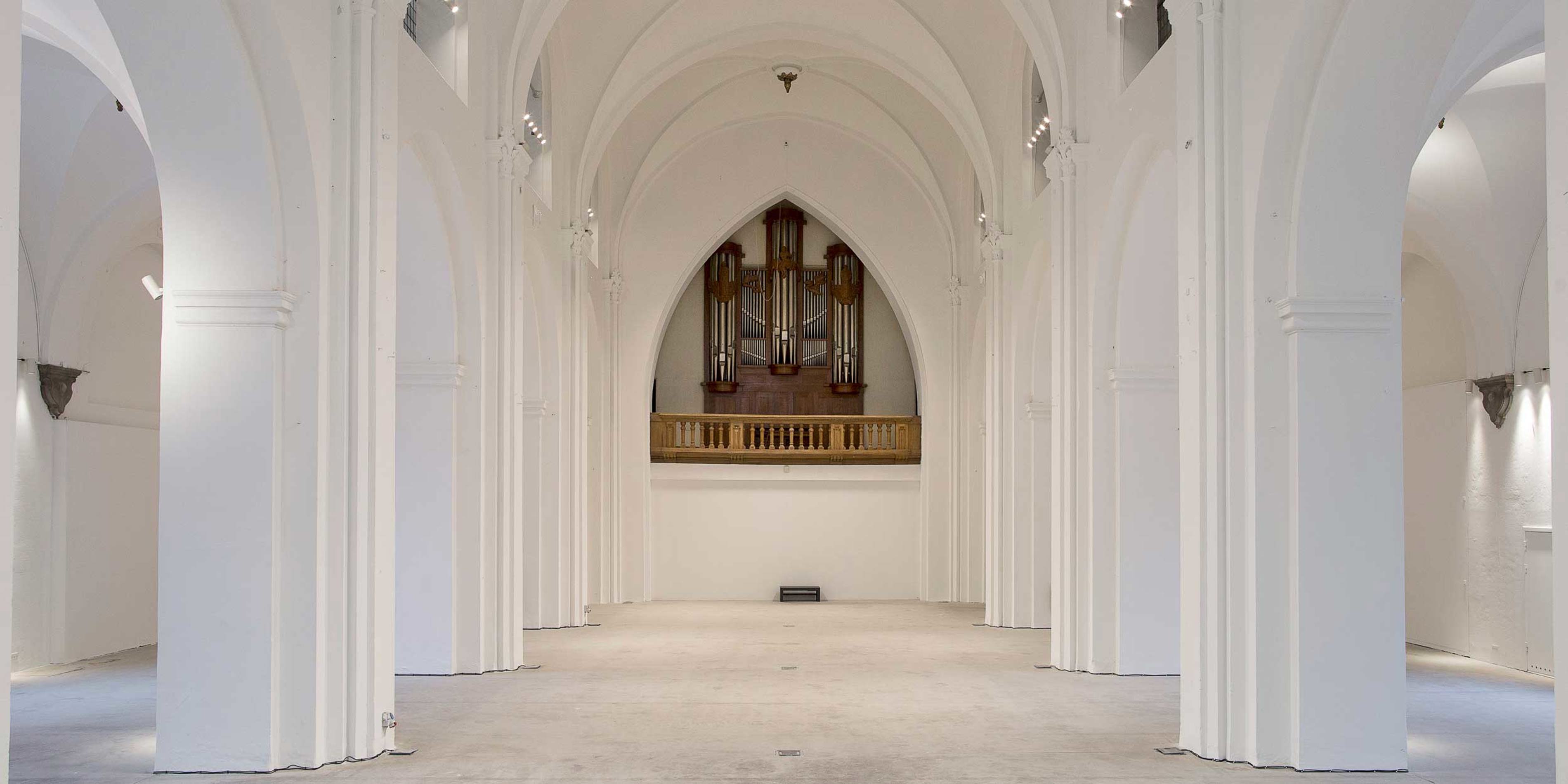 Spacious white church interior with high arched columns and an impressive organ in the center in a minimalist style.