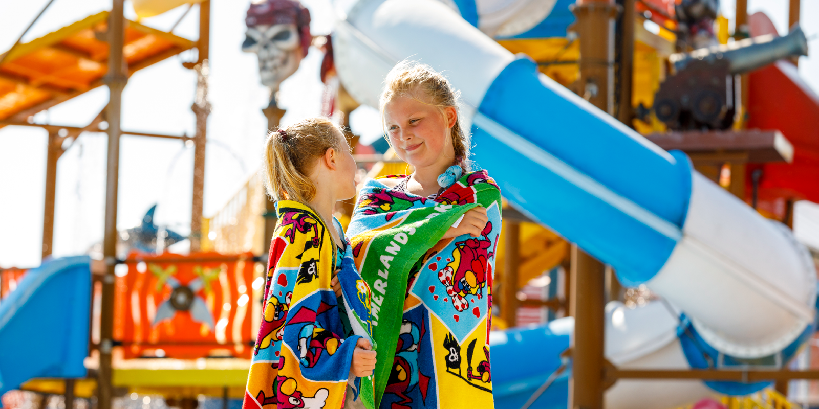 Two girls in colorful bath towels stand in front of a waterslide at a water park.