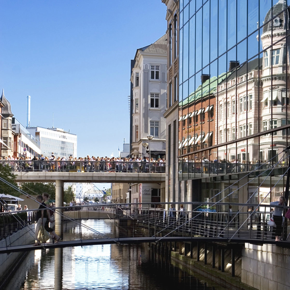 Pedestrian bridge filled with people over the stream in Aarhus city center on a sunny day, reflecting in glass buildings.