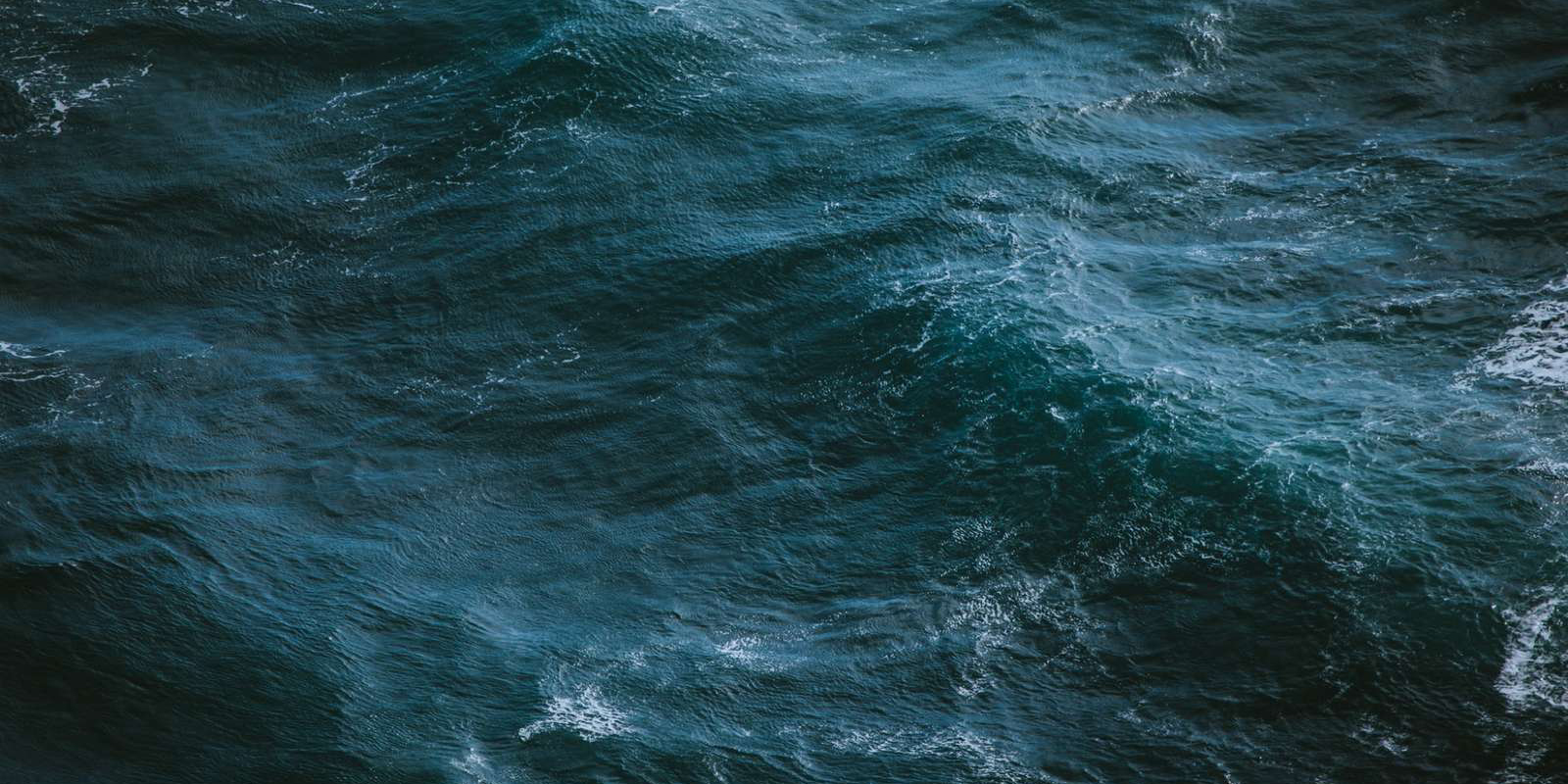 Aerial view of ocean waves with deep blue and green hues in the turbulent water.